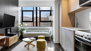 A modern kitchen with a built-in oven and microwave, a grey sofa, and a large window offering a city view. - The Cove Hotel (Montréal)
