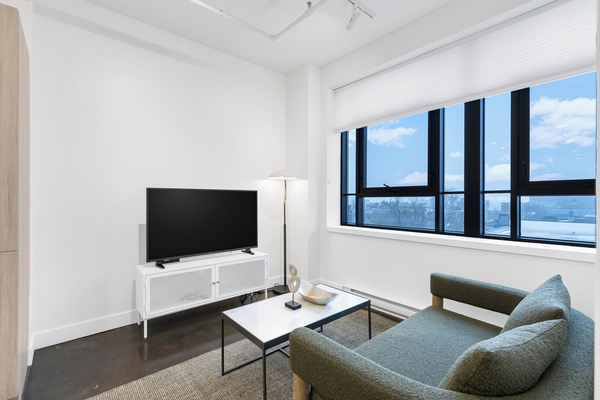 Un soggiorno moderno con una TV a schermo piatto, un mobile bianco, una lampada da terra e un'ampia finestra con vista sulla città.