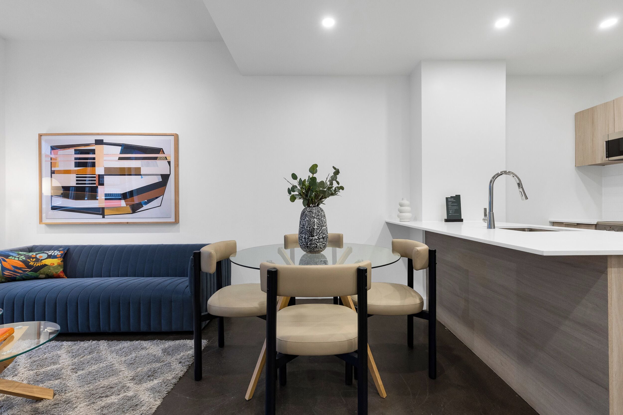 A modern living room with a blue sofa, a glass coffee table, and a dining area with four chairs.