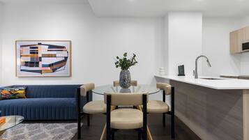 A modern living room with a blue sofa, a glass coffee table, and a dining area with four chairs.