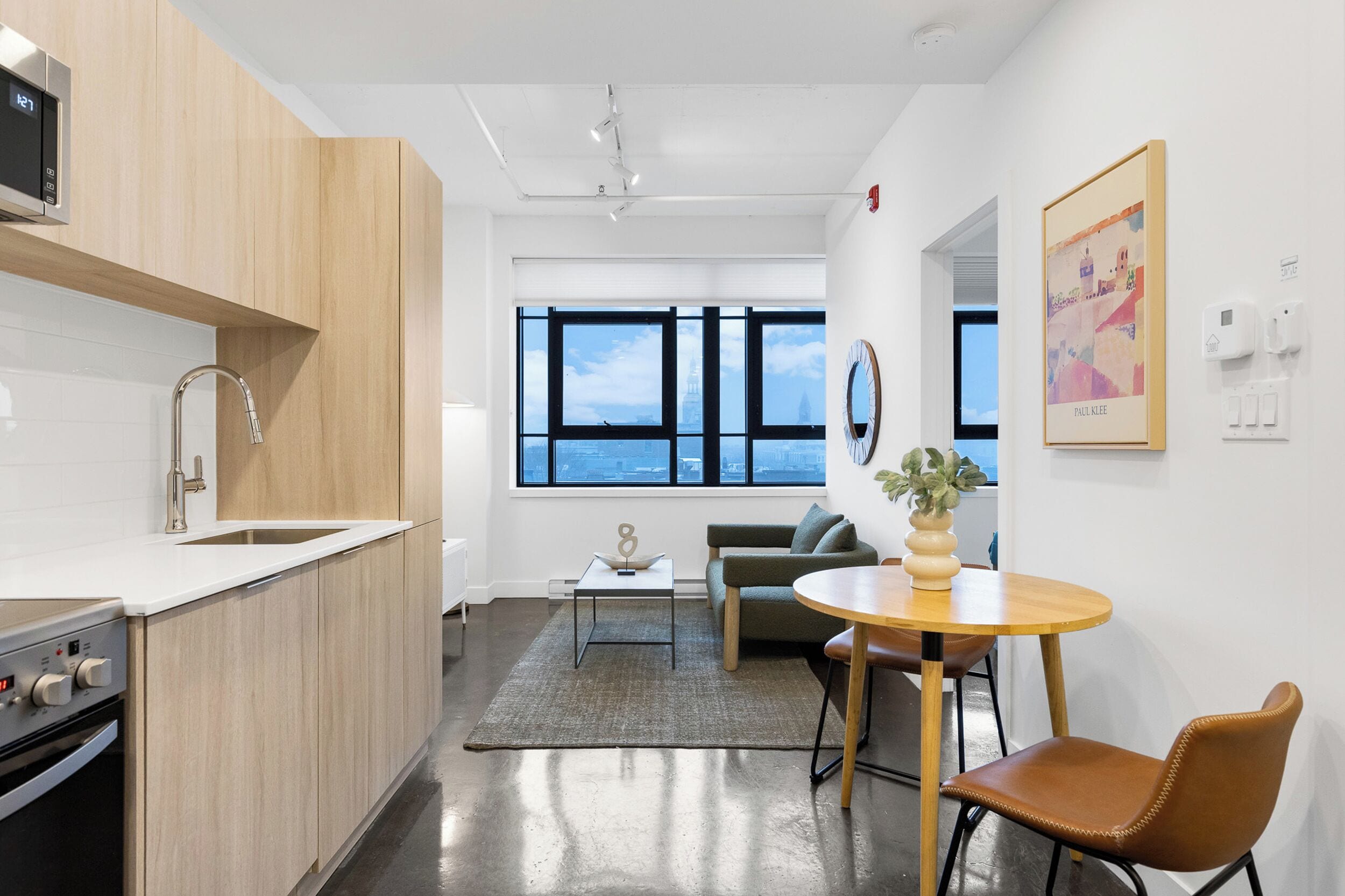 Una cocina moderna con microondas integrado, un lavamanos de acero inoxidable y encimera de madera. Cuenta con una zona de comedor con una mesa redonda y sillas, un sofá y un amplio ventanal con vistas a la ciudad.