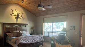 A bedroom with a wooden ceiling, a bed with a headboard, a nightstand, a lamp, a window with a view of trees, and a stuffed animal on a chair.