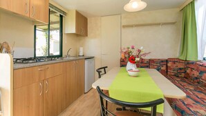 Eine kleine Küche mit Holzausstattung, einem Herd und einem Spülbecken. Ein Esstisch mit grüner Tischdecke und eine Blumenvase stehen bereit. Ein Sofa mit buntem Muster lehnt an der Wand.