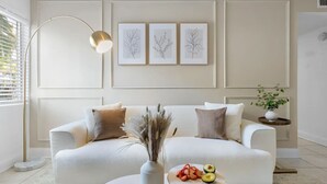 A modern living room with a white sofa, decorative pillows, a floor lamp, and framed botanical prints on the wall. - Cozy King Suite Near Bayshore | Pool & Gym (Tampa)
