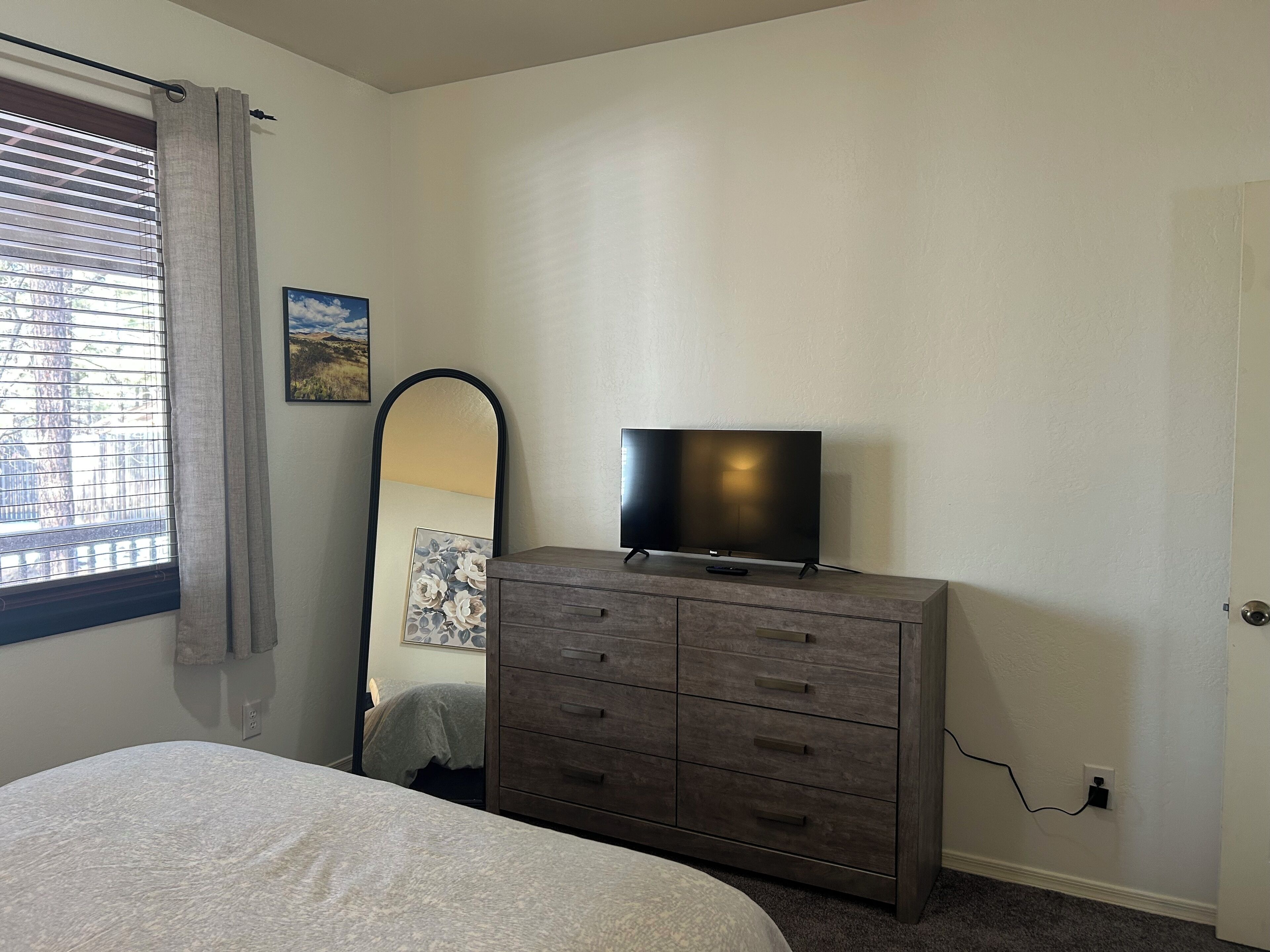 Une chambre à coucher comprenant un lit, une commode sur laquelle est posée une télévision, une fenêtre avec des stores et un miroir fixé au mur.
