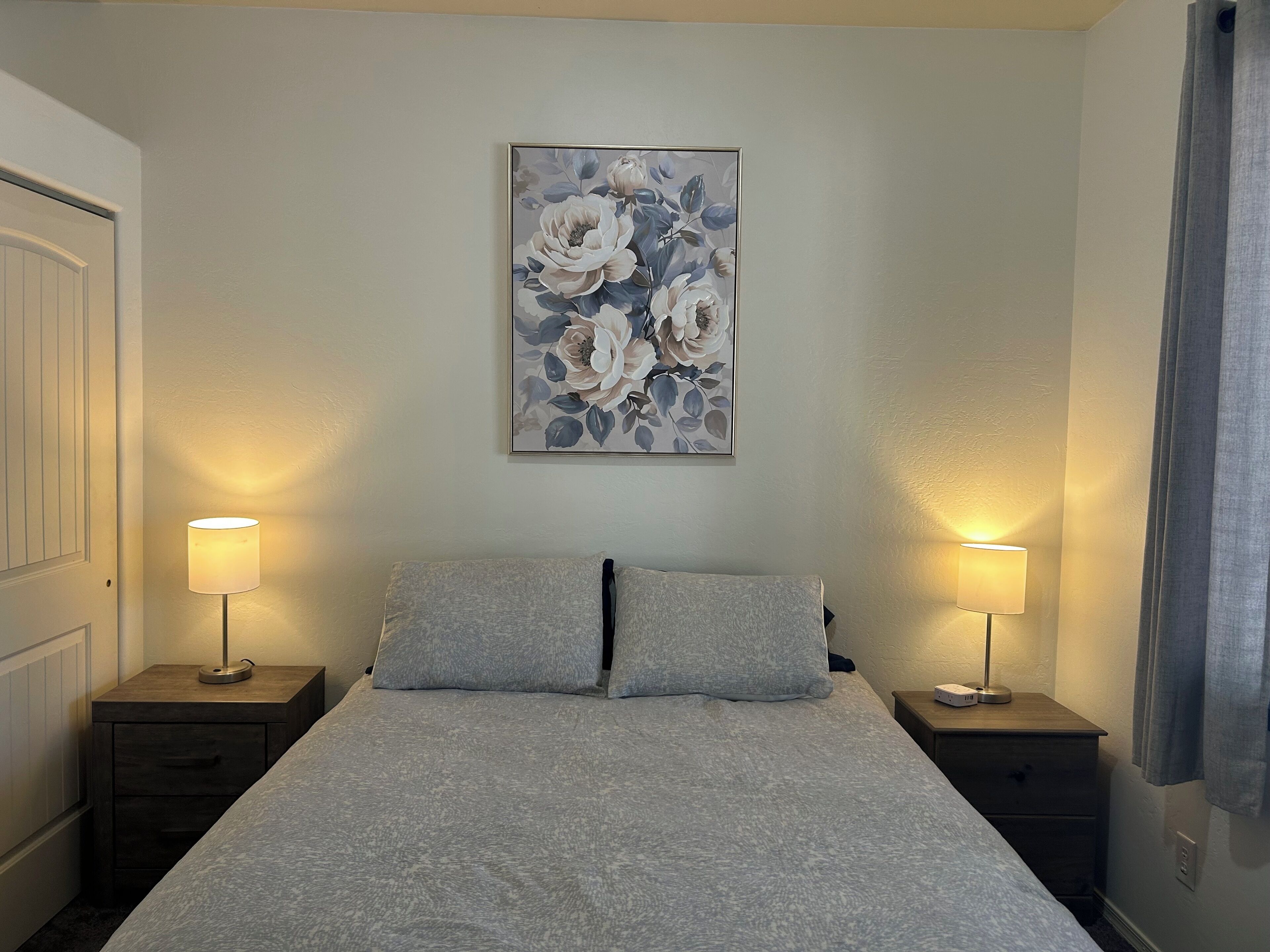 A bedroom with a bed, two bedside tables with lamps, a wall-mounted painting, and a door.