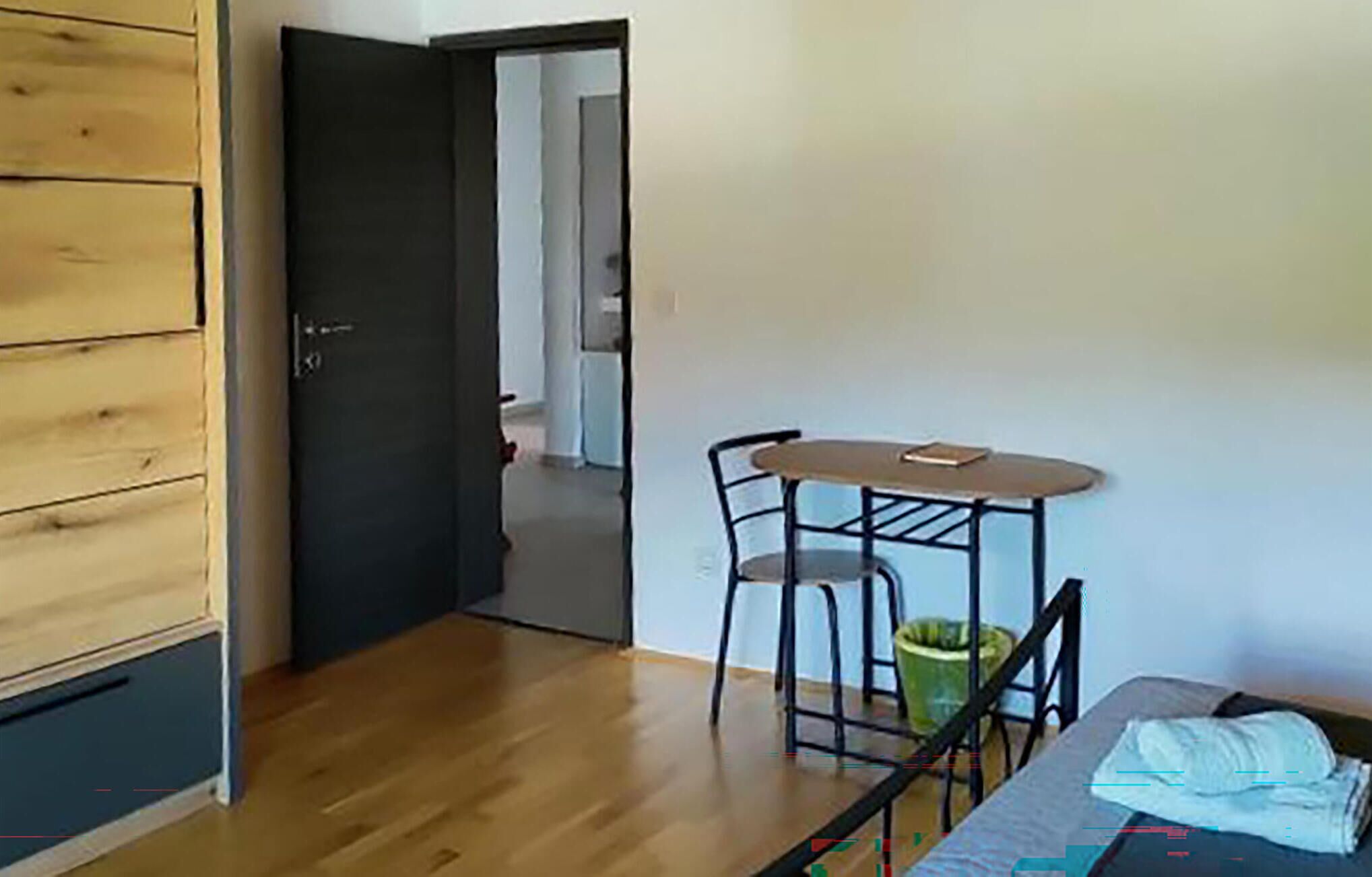Une pièce avec une porte en bois, une petite table avec deux chaises et un lit, à côté duquel se trouve une poubelle verte.