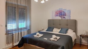 Cama bem arrumada com cabeceira cinza, edredom azul e branco e duas toalhas dobradas sobre a cama. Tem uma mesinha de cabeceira com abajur, uma janela com persianas e um quadro de uma árvore na parede.