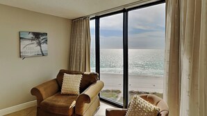 Een kamer met een groot raam dat uitkijkt op een strand, een bruine bank en een fauteuil.