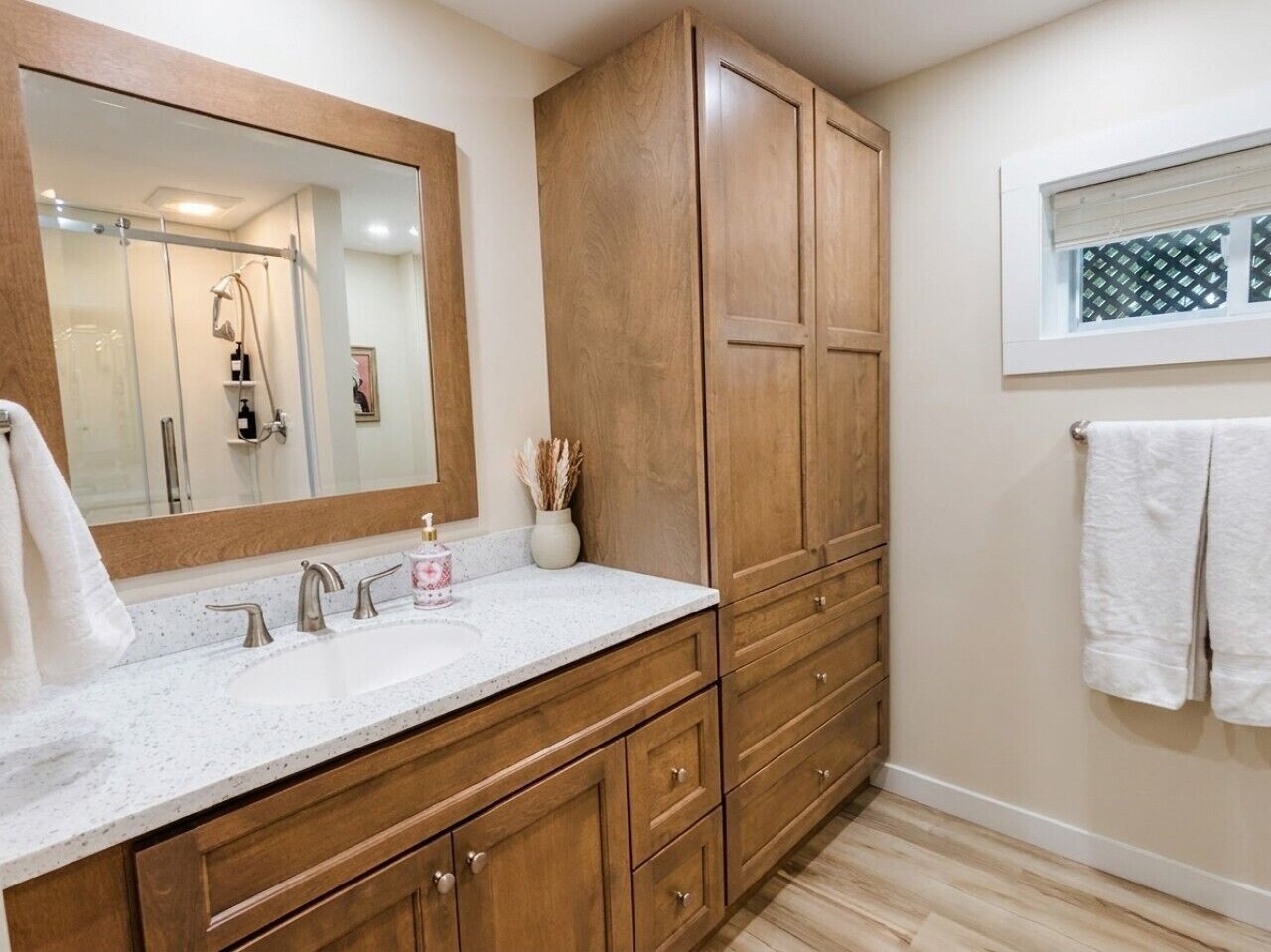 A bathroom with a large mirror, wooden cabinets, a white countertop, and a window with blinds.