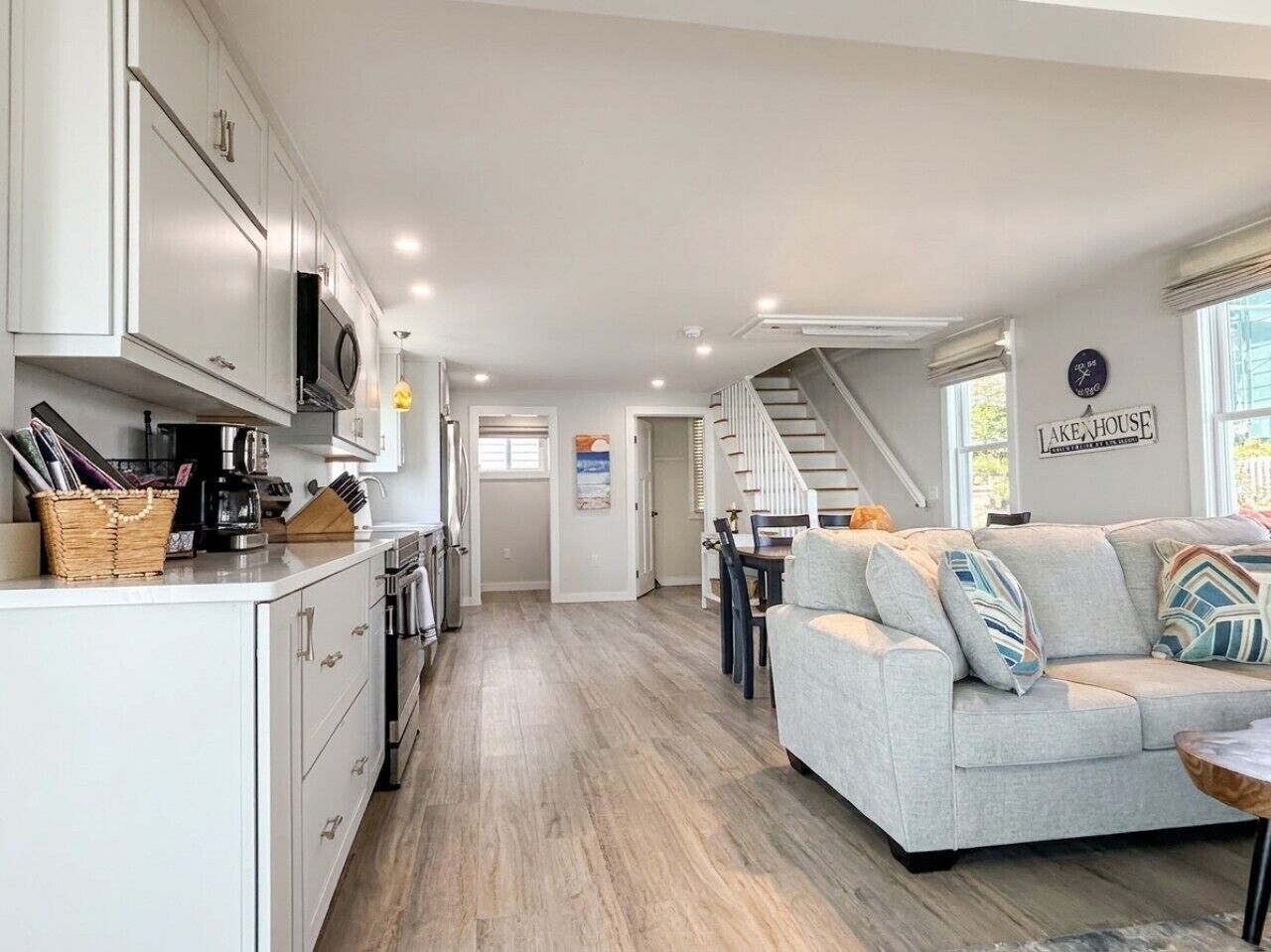 A modern kitchen with white cabinets, a microwave, and a coffee maker. There is a dining area with a gray sofa and patterned cushions. A staircase leads to an upper level.