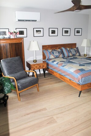 Ein Schlafzimmer mit einem hölzernen Bett, einem grauen Sessel, einem Nachttisch aus Holz, einer Wand-Klimaanlage und gerahmten Bildern an der Wand.