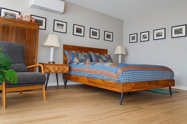 Ein Schlafzimmer mit einem hölzernen Bett, einem grauen Sessel, einem Nachttisch aus Holz und einer Topfpflanze.