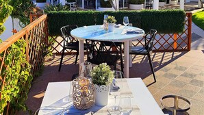 Un área de comedor al aire libre con una mesa dispuesta para dos, rodeada de sillas, y vista a un edificio con fachada blanca y ventanas grandes.