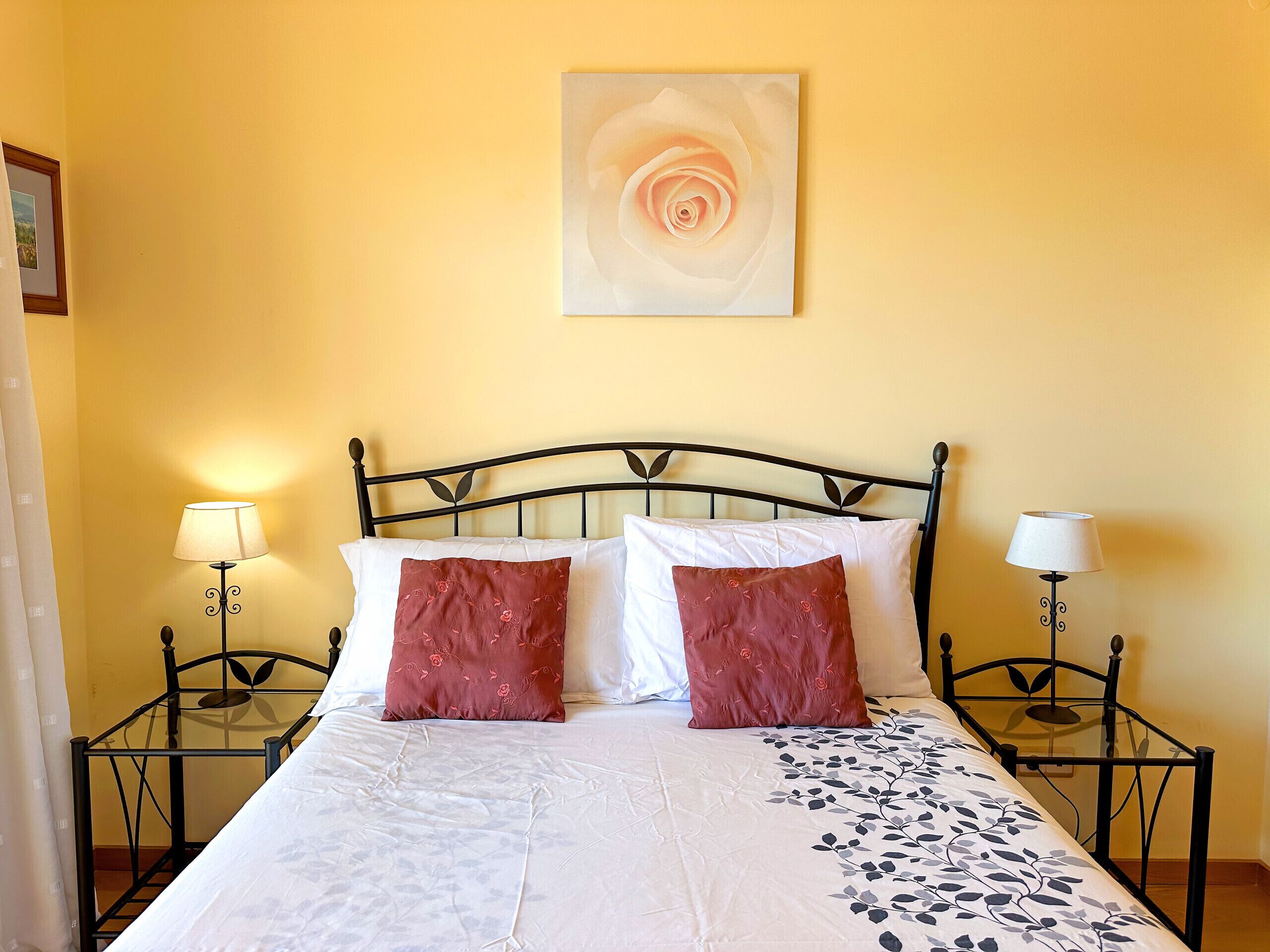 Una cama bien tendida con almohadas blancas y rojas, un edredón estampado con flores y dos mesitas de noche con lámparas.
