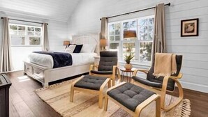 Un dormitorio con cama, dos sillones, una mesita de noche y una ventana con cortinas.