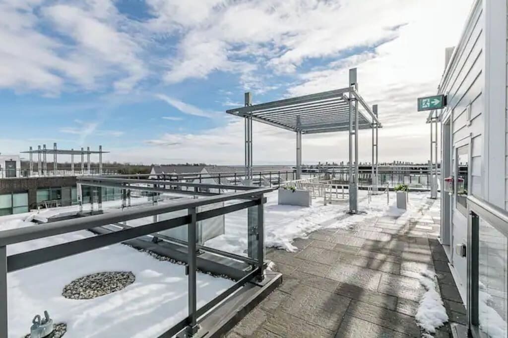 Terraço no telhado com guarda-corpo de vidro, pérgola de metal e chão coberto de neve.