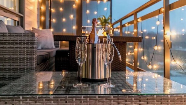 Une table en verre avec un seau à champagne et des verres, sur un balcon illuminé de guirlandes lumineuses.