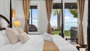 Ein ordentlich bezogenes Bett mit einer beige farbenen Tagesdecke, Blick auf das Meer durch große Fenster und eine Lampe auf dem Nachttisch.