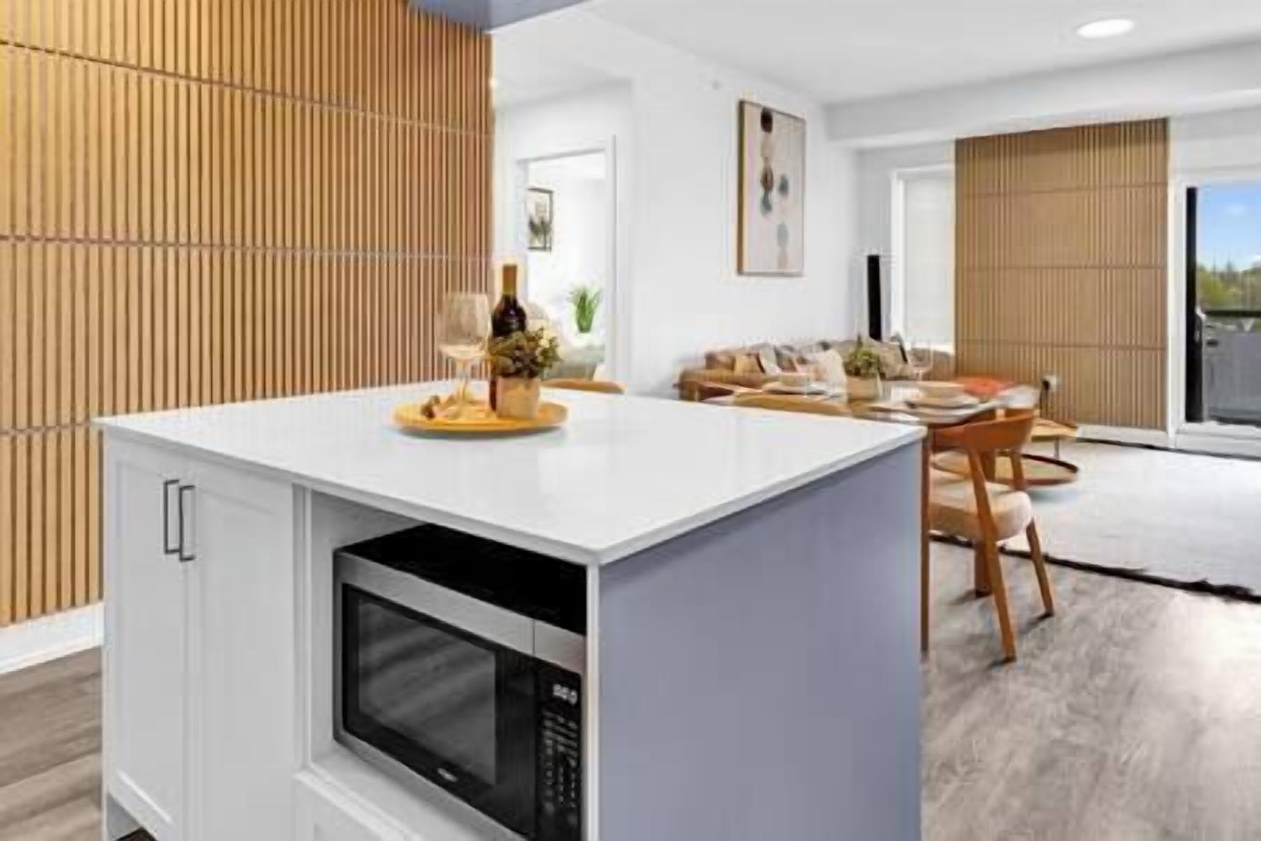 Uma cozinha moderna com micro-ondas embutido, sala de jantar com mesa e cadeiras de madeira, e sala de estar com sofá e uma janela grande.