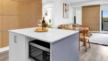 Uma cozinha moderna com micro-ondas embutido, sala de jantar com mesa e cadeiras de madeira, e sala de estar com sofá e uma janela grande.