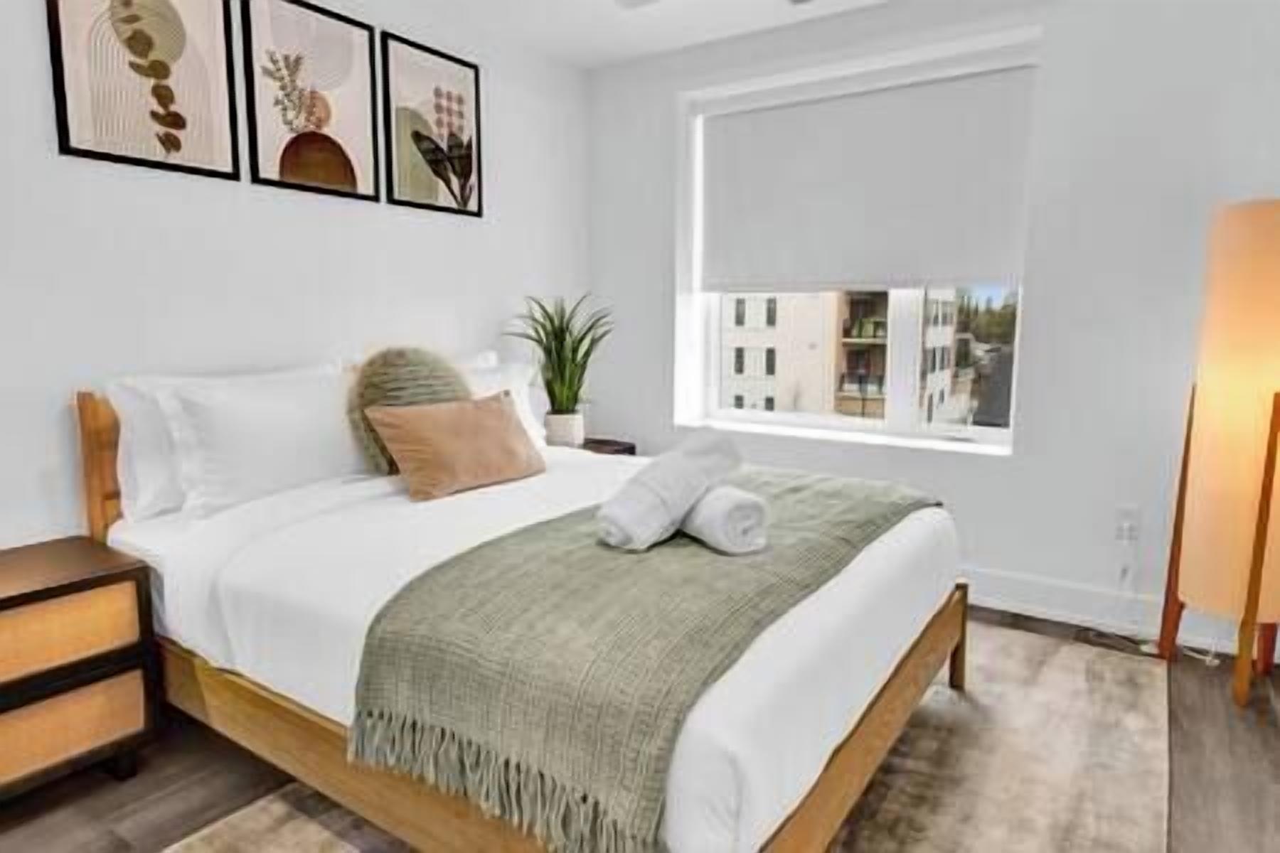 A bedroom with a bed, pillows, a blanket, a nightstand, a lamp, and a window with a view of buildings.