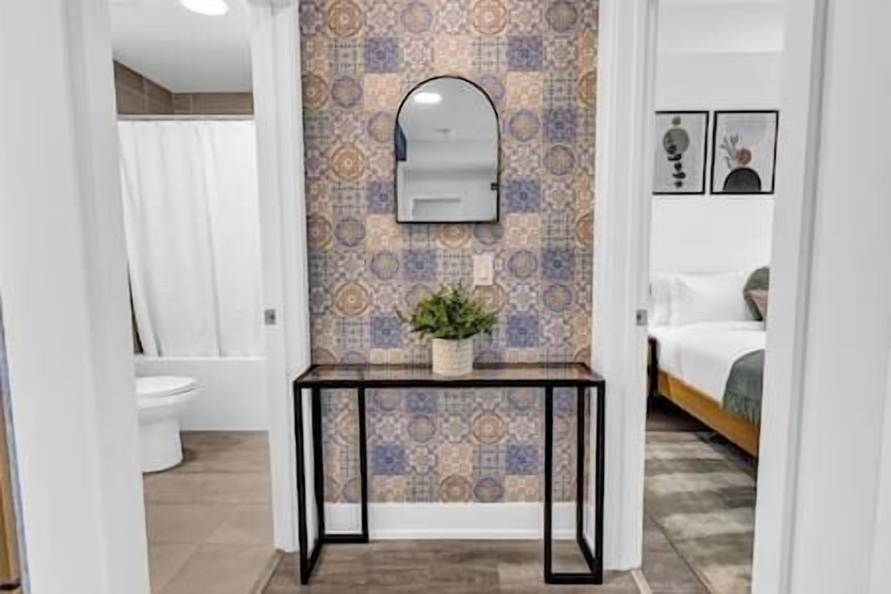 A bathroom with a patterned tile wall, a mirror above a console table, and a toilet visible in the background.