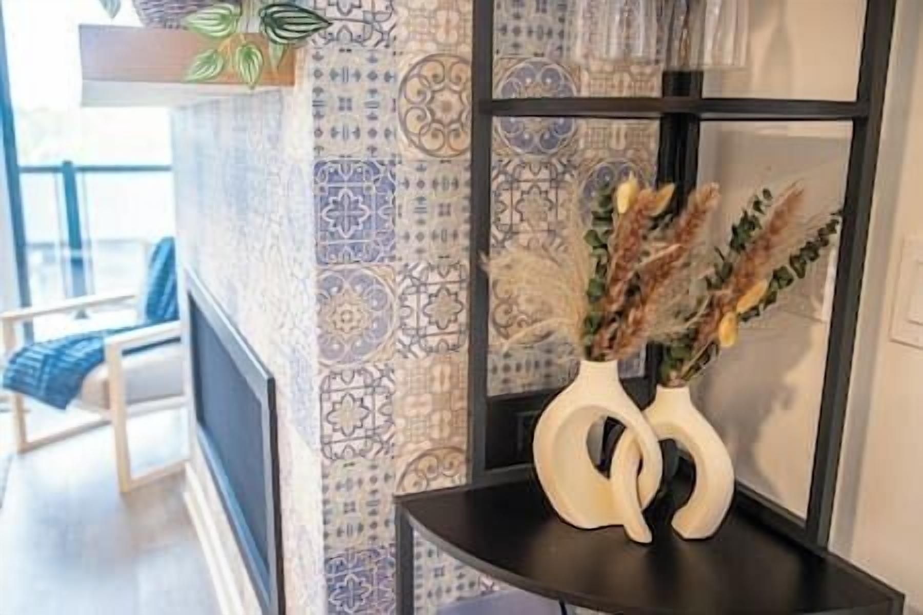 A room with patterned wallpaper, a vase with dried plants, and a chair with a blue cushion.