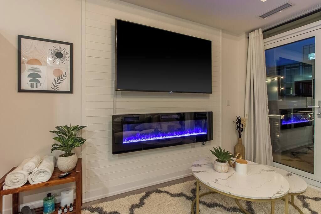 A modern living room with a flat-screen TV mounted above a fireplace, a small round table with a vase, and a large window with a view of the city.