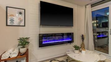 A modern living room with a flat-screen TV mounted above a fireplace, a small round table with a vase, and a large window with a view of the city.