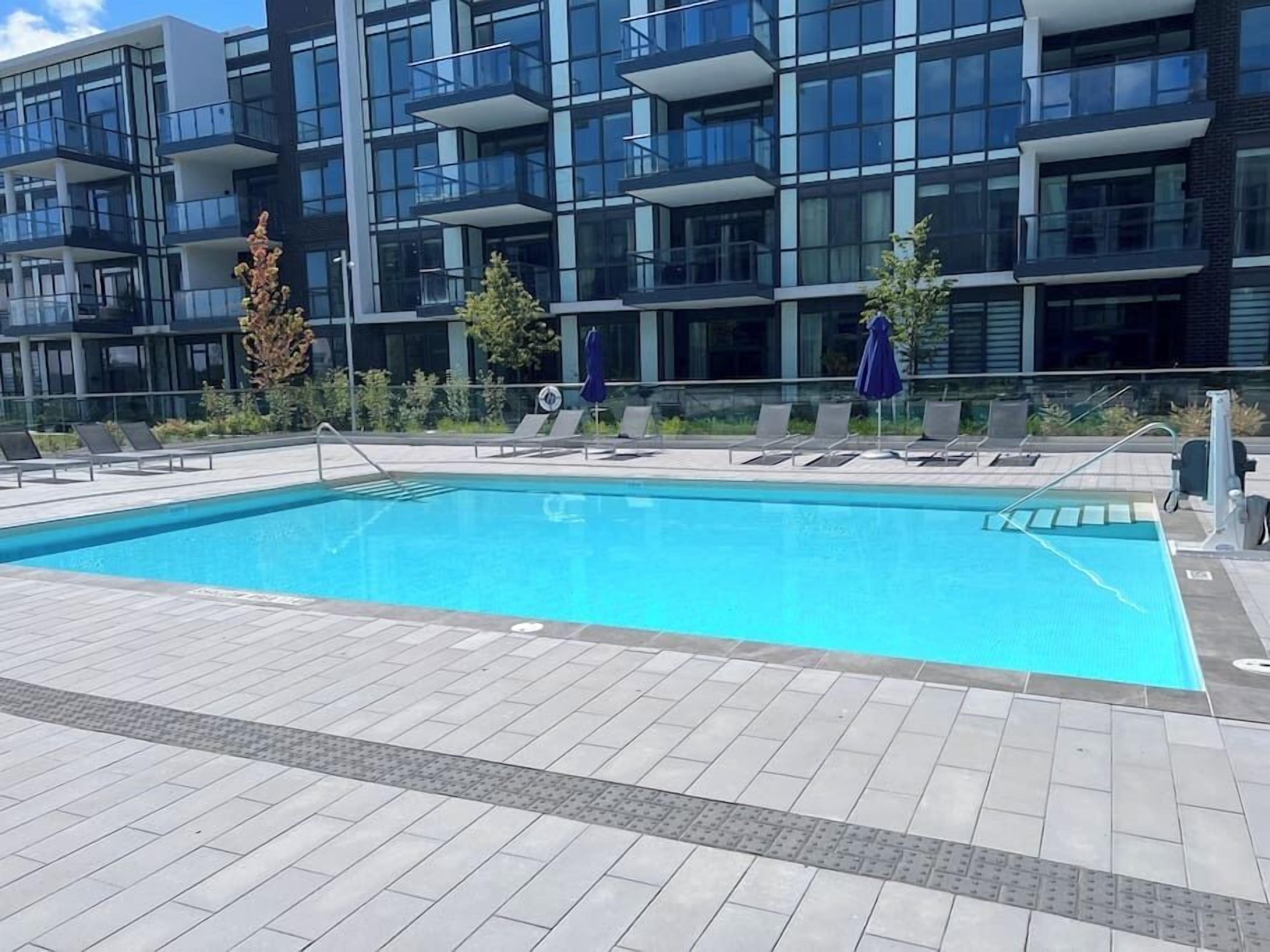 A modern building with a swimming pool, sun loungers, and a blue umbrella.