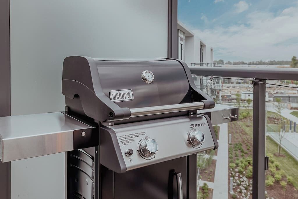 Un grill Weber Spirit amb dues manetes de control, col·locat en una barana de balcó amb vista a la ciutat.