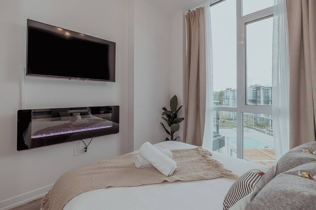 Een moderne hotelkamer met een groot bed, een tv aan de muur, een klein tafeltje met een plant en uitzicht op een stadsbeeld door een groot raam.