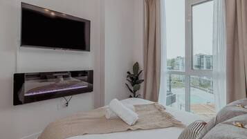 Een moderne hotelkamer met een groot bed, een tv aan de muur, een klein tafeltje met een plant en uitzicht op een stadsbeeld door een groot raam.