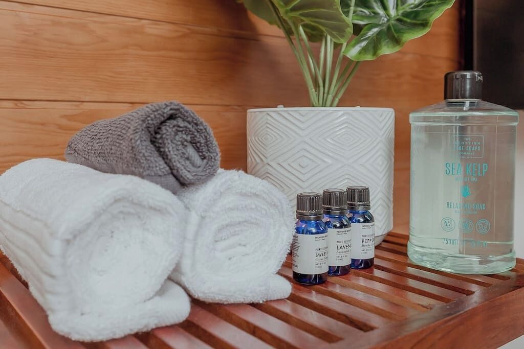 A wooden tray with a white towel, a rolled gray towel, and three small blue bottles labeled with scents.