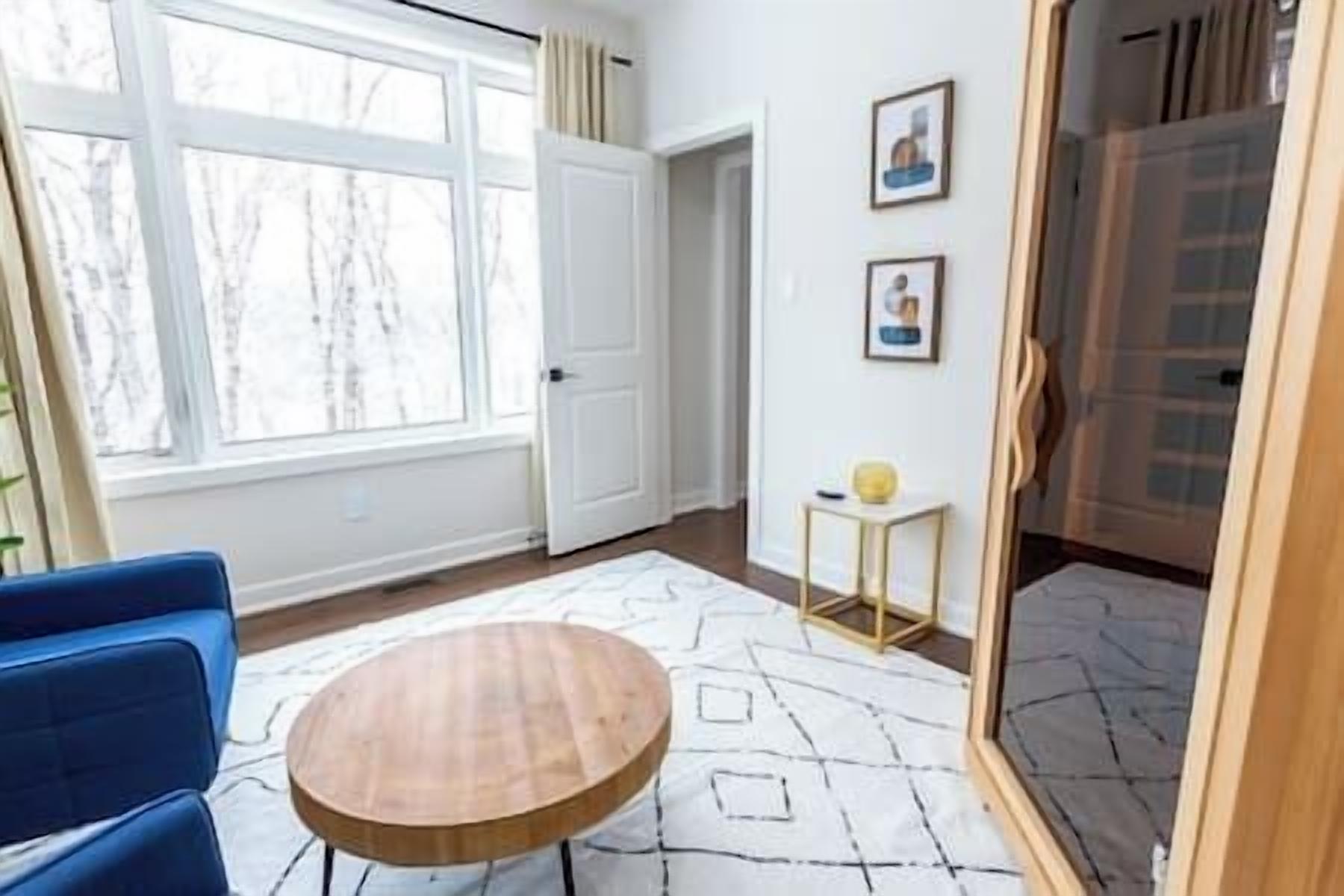 A room with a large window, a round wooden table, a blue chair, and a small side table with a lamp.
