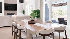 A modern dining area with a long wooden table and white chairs, a potted plant, and a large window offering a view of the outdoors. - The Algonquin Lakehouse w Hot Tub, Games, Fire Pit (Huntsville)