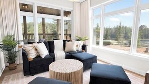 A modern living room with a sectional sofa, round ottoman, and large windows offering a view of the outdoors. - The Algonquin Lakehouse w Hot Tub, Games, Fire Pit (Huntsville)