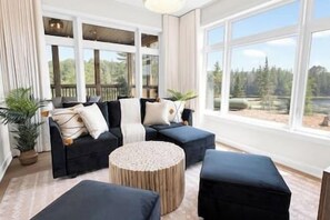 Ein modernes Wohnzimmer mit Ecksofa, rundem Hocker und großen Fenstern mit Blick ins Freie.