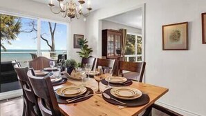 Ein Esszimmer mit Holztisch, gedeckt für sechs Personen, Stühlen, einer Kristallleuchtre und Meerblick durch große Fenster.