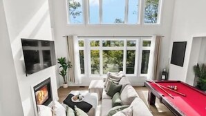 A spacious living room with a red pool table, a comfortable sofa set, and a large window offering a view of greenery. - Northern Lights Muskoka w/ Hot Tub & Pool Table (Muskoka District Municipality)