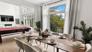 A dining area with a long table set for six, surrounded by chairs. There is a pool table in the background, and the room has large windows offering a view of greenery outside. - Northern Lights Muskoka w/ Hot Tub & Pool Table (Muskoka District Municipality)