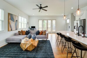 Ein großzügiger Wohnbereich mit grauem Sofa, einem Holztisch und einem schwarzen Teppich mit Muster. Drei Barhocker und eine Küche mit einem Edelstahlkühlschrank sind ebenfalls vorhanden.
