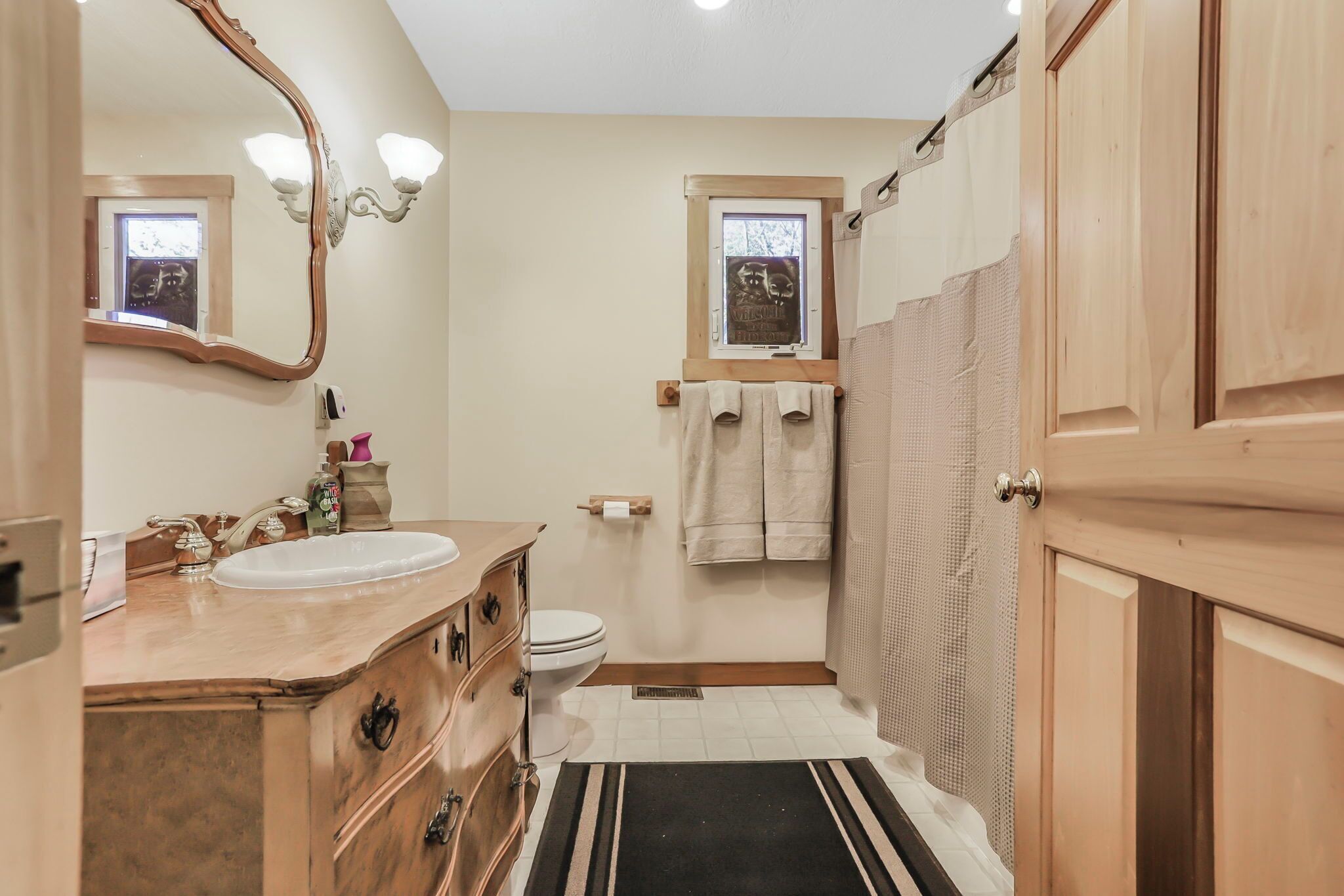 Een badkamer met een houten wastafelmeubel, een wastafel, een toilet, een douchegedeelte en een raam met een handdoek aan de muur.