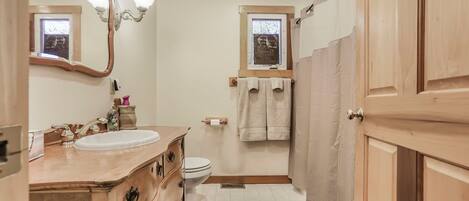 Een badkamer met een houten wastafelmeubel, een wastafel, een toilet, een douchegedeelte en een raam met een handdoek aan de muur.