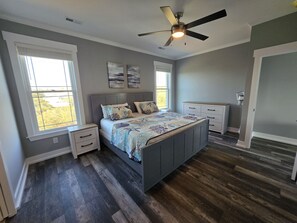 A bedroom with a large bed, two bedside tables, a ceiling fan, and two windows. - Spacious Beach Home for Large Groups • Sleeps 14 • Walk to Access (Oak Island)