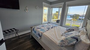 Une chambre à coucher comprenant un lit, un ventilateur de plafond, un téléviseur fixé au mur et de grandes fenêtres donnant sur l’extérieur.