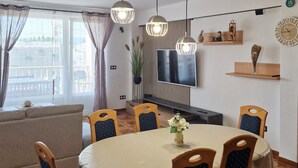 A modern living room with a round dining table, wooden chairs, a flat-screen TV, and a large window with curtains. - Amazing apartment in Omis (Omis)