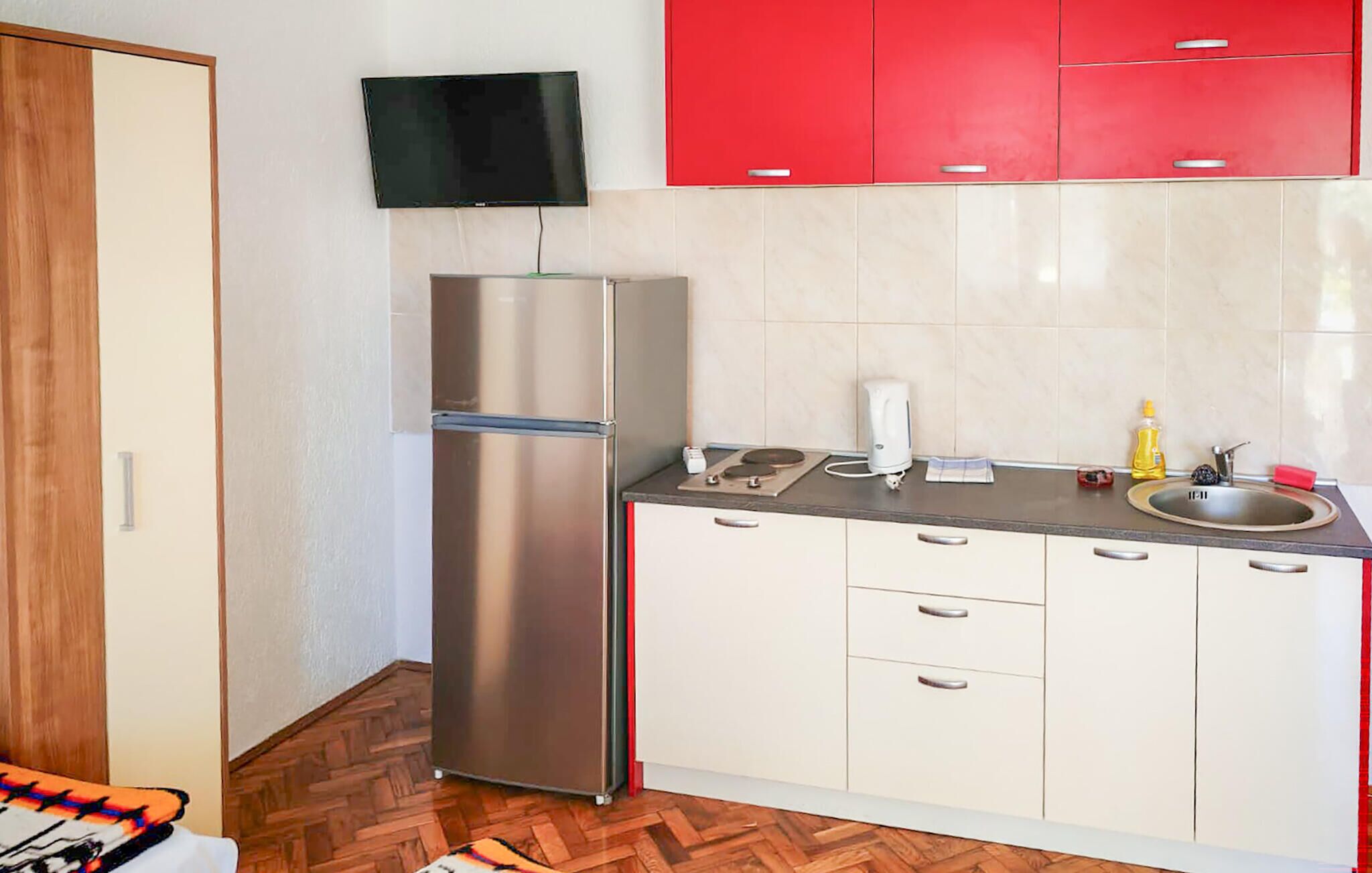 Una cucina compatta con un frigorifero in acciaio inossidabile, una TV a schermo piatto montata sopra e un lavello con un pensile rosso.