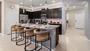 Cozinha moderna com armários escuros, uma ilha central com banquetas, eletrodomésticos de aço inoxidável e iluminação pendente.